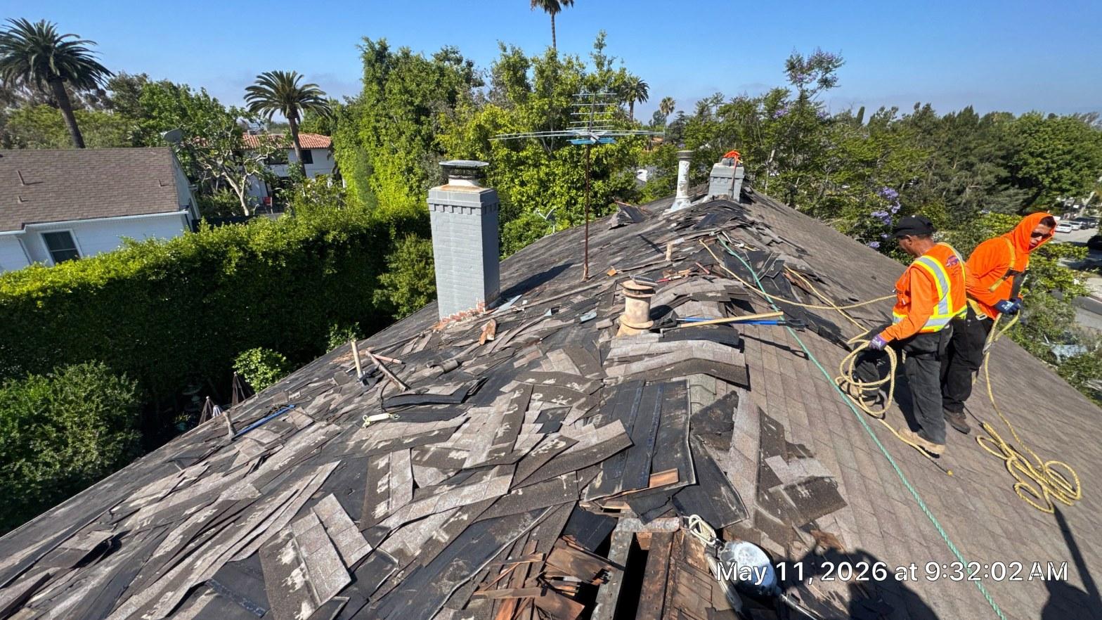 shingle-reroof-motor-ave-los-angeles-611 Two workers in orange safety vests tearing off worn asphalt shingles on steep roof with chimneys visible.