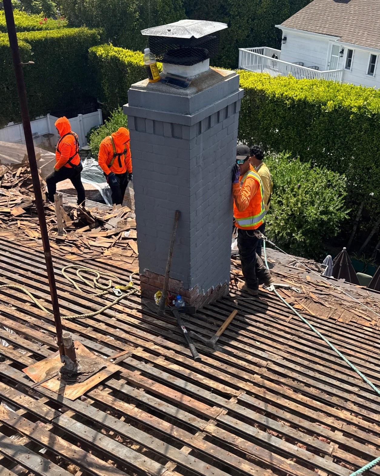 shingle-reroof-motor-ave-los-angeles-612 Roofers in orange safety vests stripping clay tiles from wood lath roof deck around gray painted chimney.