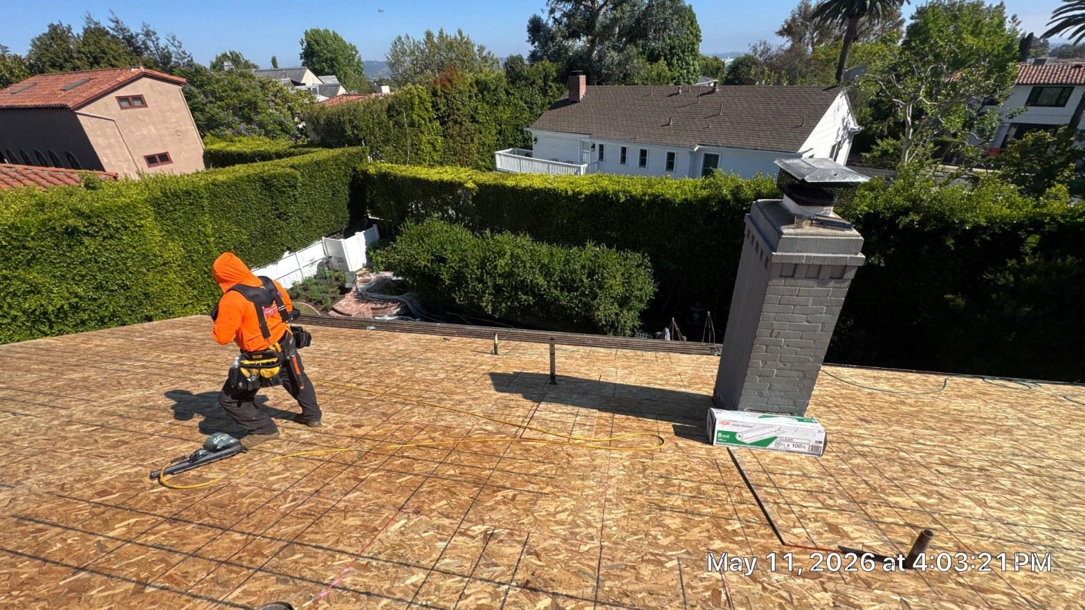 shingle-reroof-motor-ave-los-angeles-617 Worker in orange safety gear on OSB roof decking beside brick chimney, roofing materials and nail gun visible.