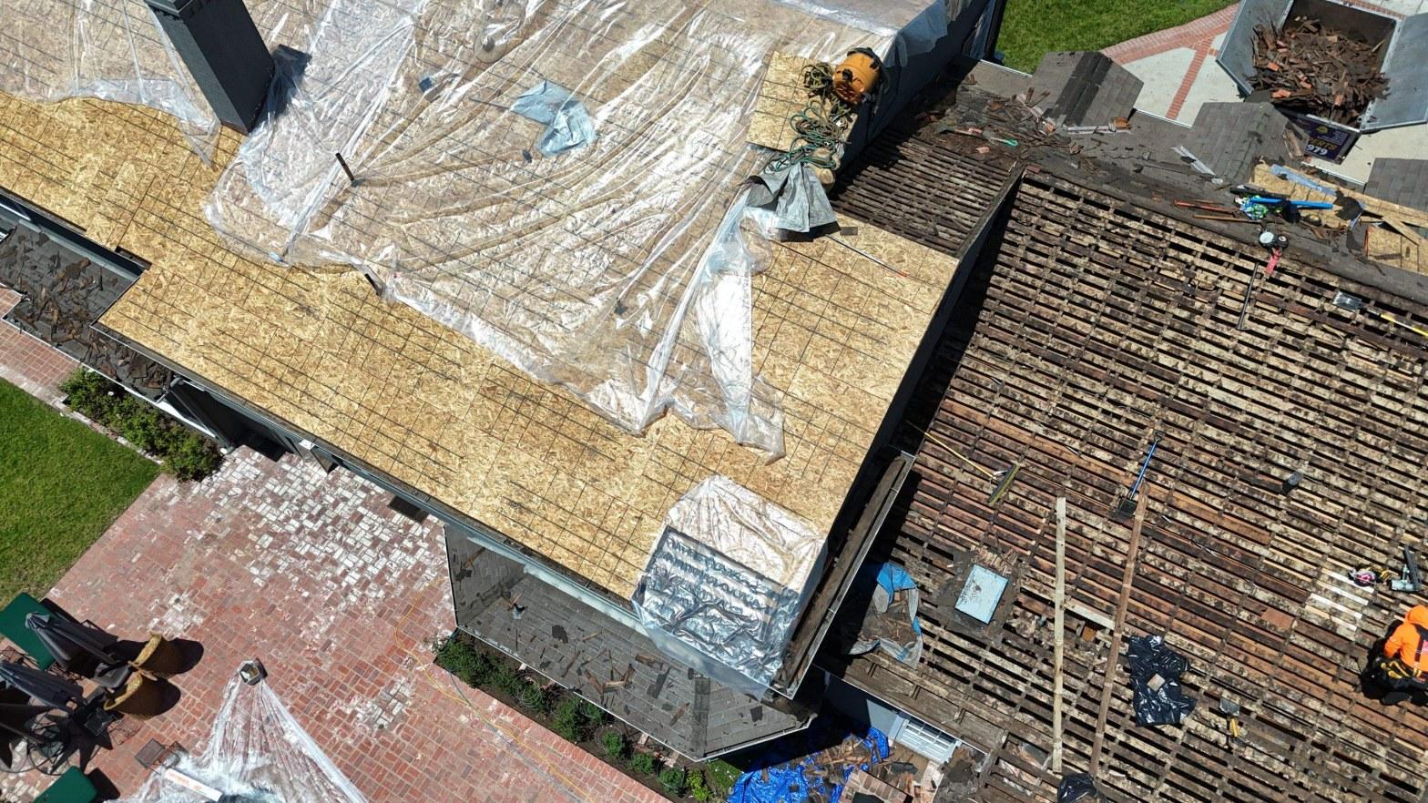 shingle-reroof-motor-ave-los-angeles-618 Aerial view of roof replacement showing OSB decking, plastic sheeting, stripped tile battens, and worker in orange vest.
