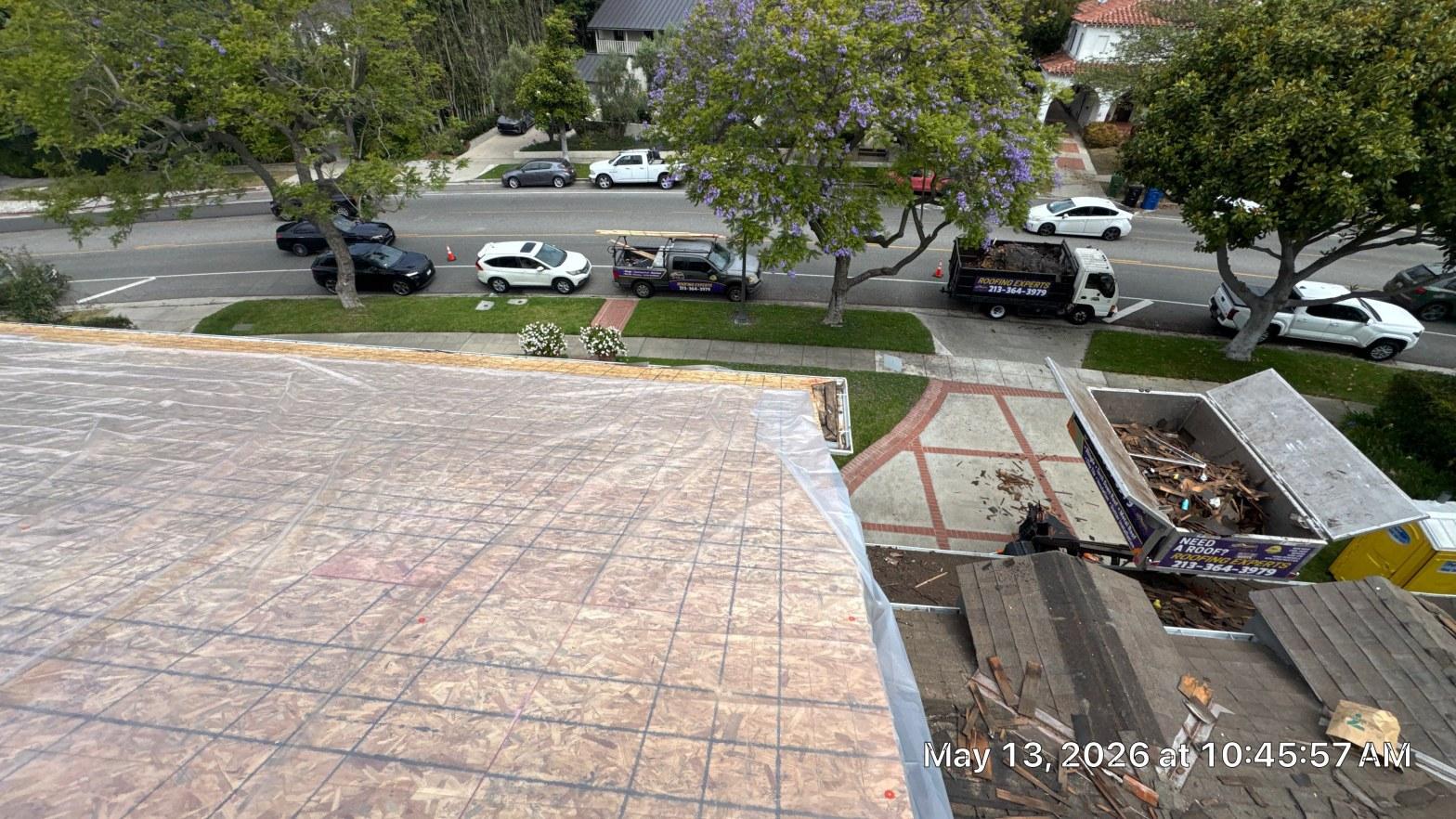 shingle-reroof-motor-ave-los-angeles-620 Aerial view of stripped roof deck covered in underlayment, dumpster filled with old roofing debris, Roofing Experts truck on street.