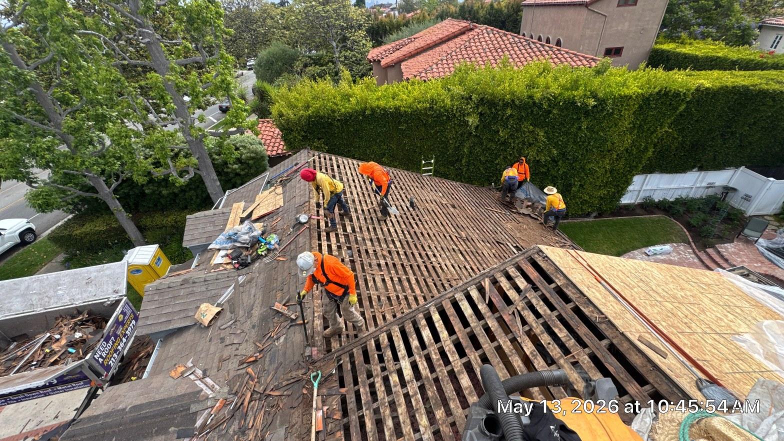 shingle-reroof-motor-ave-los-angeles-621 Roofing crew in orange safety gear tearing off wood lath strips on steep residential roof, debris visible.