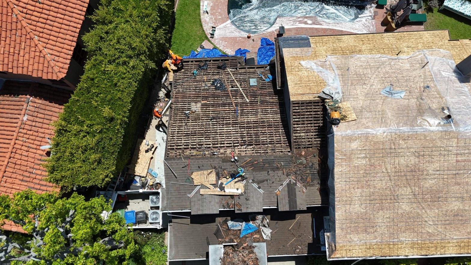 shingle-reroof-motor-ave-los-angeles-622 Drone overhead shot of residential roof replacement in progress, exposed wood battens, sheathing, debris, and two workers on site.