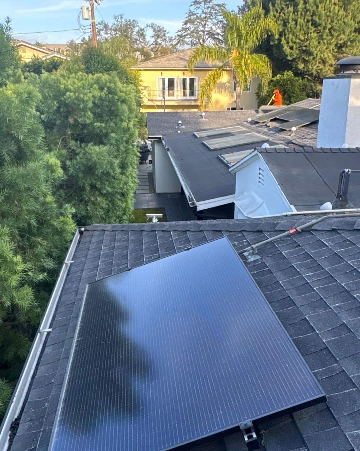 shingle-reroof-solar-complex-flashing-los-angeles-643 Black solar panels mounted on dark shingle roof, worker in orange vest on adjacent rooftop, residential neighborhood.