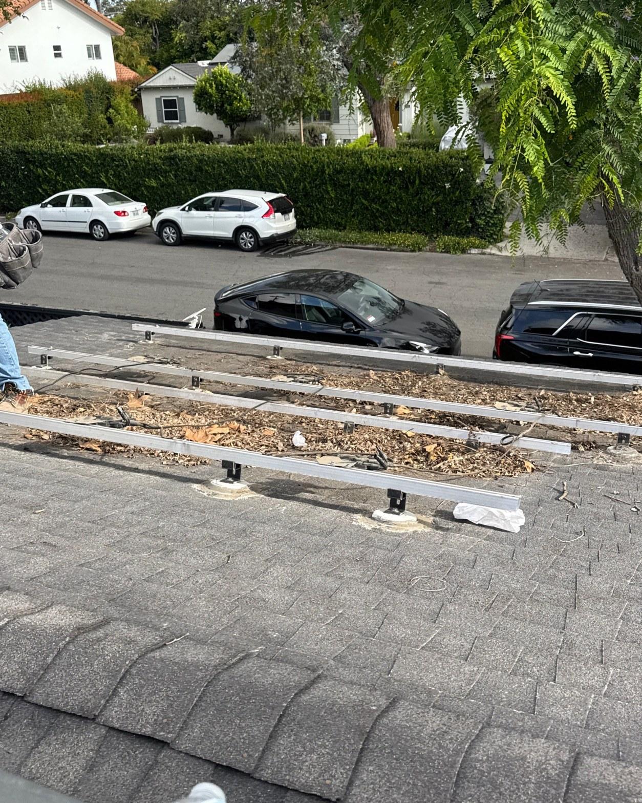 shingle-reroof-solar-complex-flashing-los-angeles-649 Aluminum solar panel racking rails bolted to gray asphalt shingle roof, leaves accumulated underneath, residential street visible below.