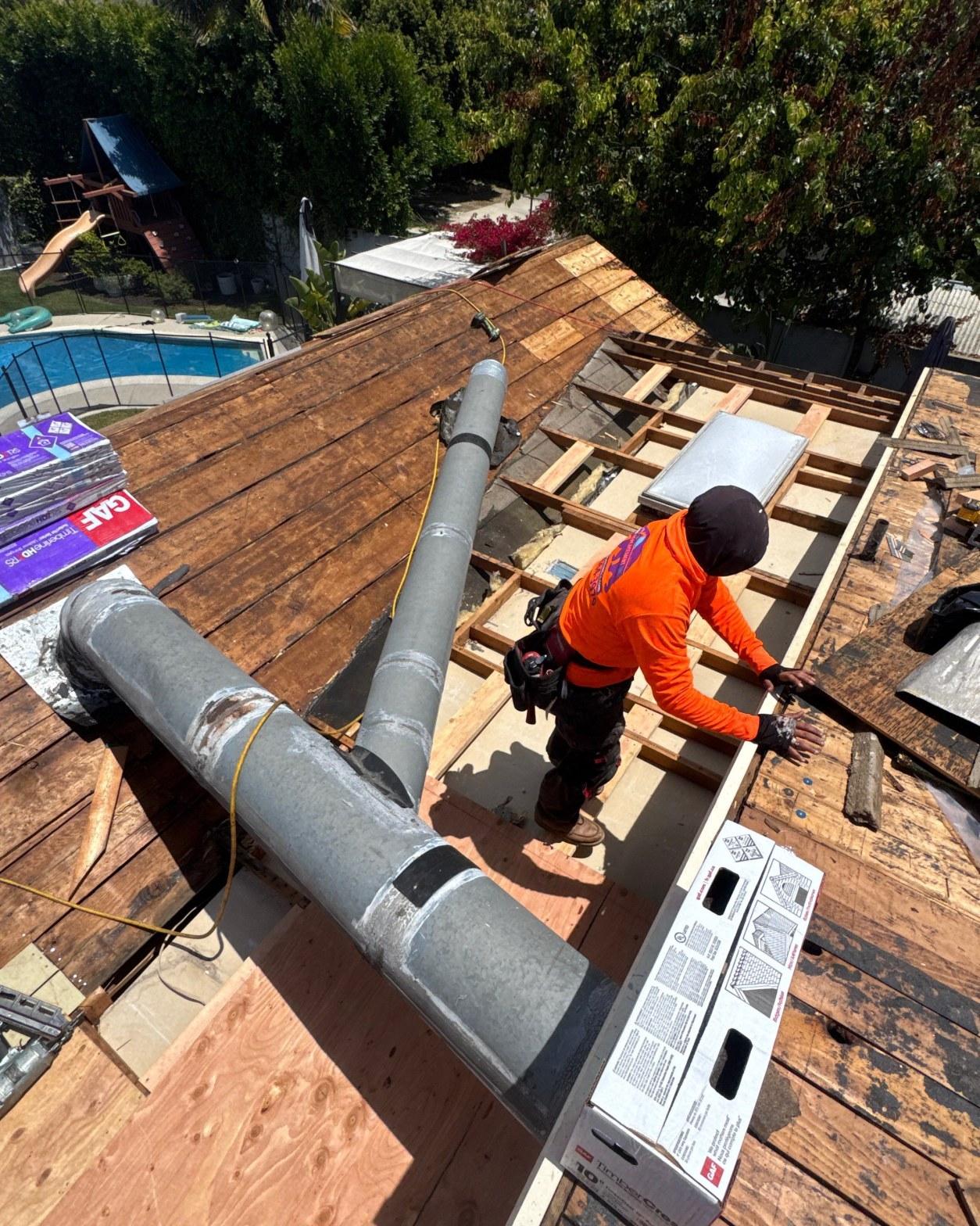 single-reroof-burbank-ca-shingles-flat-flashing-575 Roofer on stripped residential roof installing plywood decking near metal chimney flues, GAF shingles visible nearby.