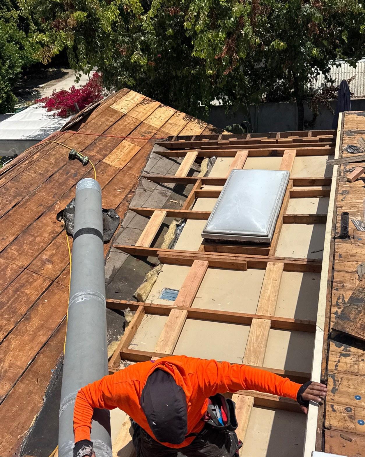 single-reroof-burbank-ca-shingles-flat-flashing-577 Roofer in orange shirt installing skylight on exposed wood roof deck with framing and vent pipe visible.