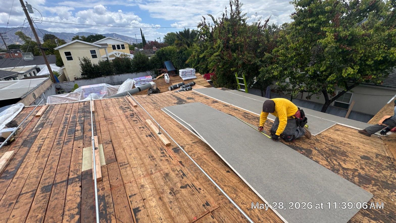 single-reroof-burbank-ca-shingles-flat-flashing-578 Roofer in yellow jacket measuring gray underlayment on flat roof wood decking during re-roofing project, Los Angeles.