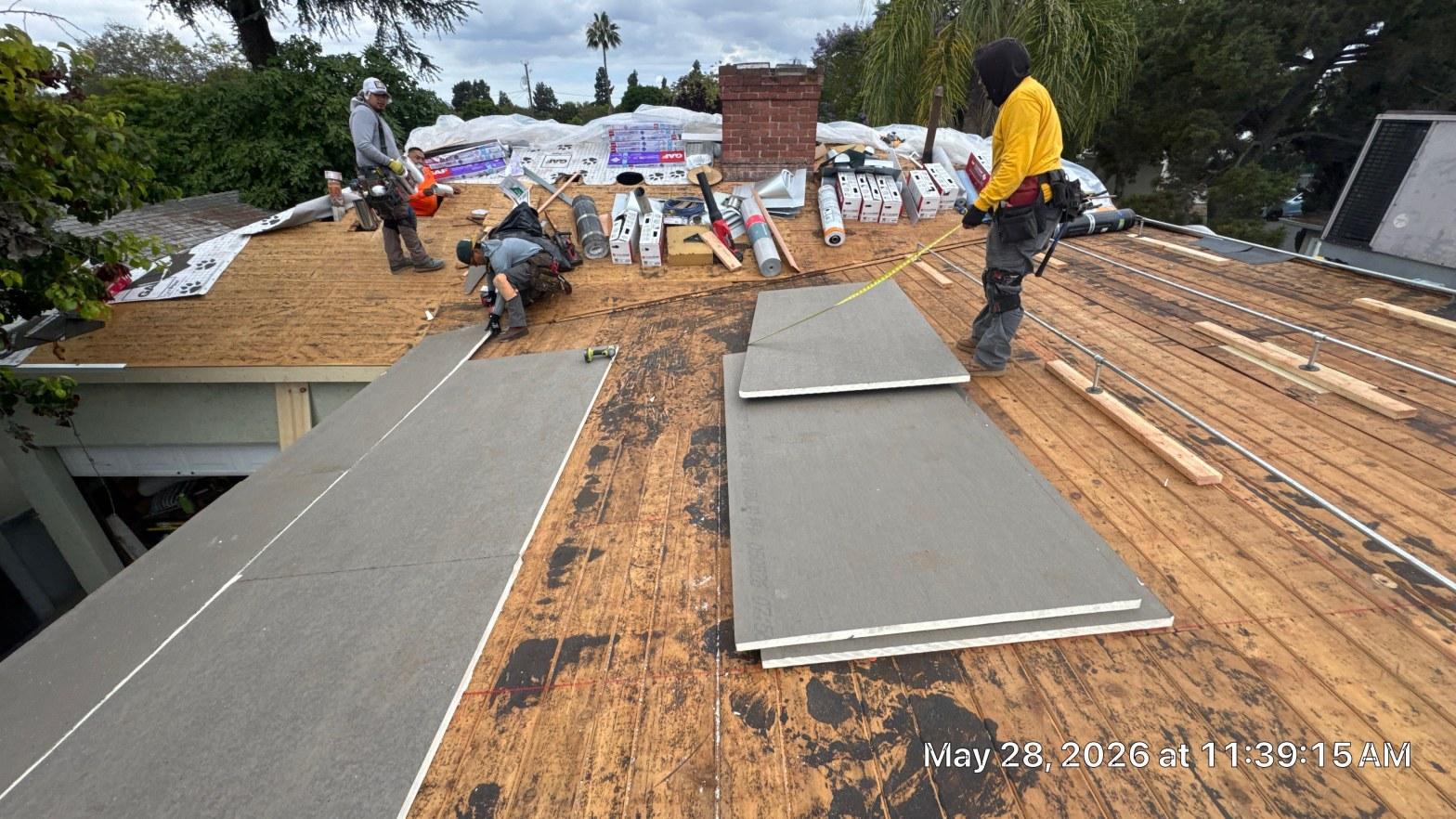 single-reroof-burbank-ca-shingles-flat-flashing-579 Three roofers laying gray underlayment and wood battens on exposed roof decking with materials and brick chimney visible.