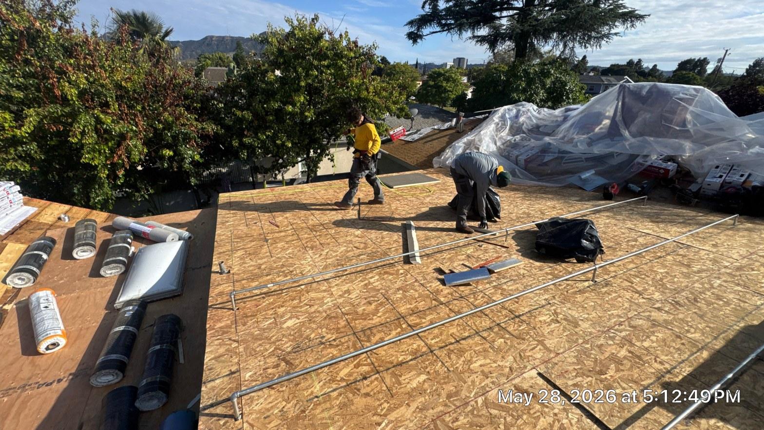 single-reroof-burbank-ca-shingles-flat-flashing-580 Two workers on flat roof with OSB decking, roofing rolls and tools, Hollywood Hills visible in background.