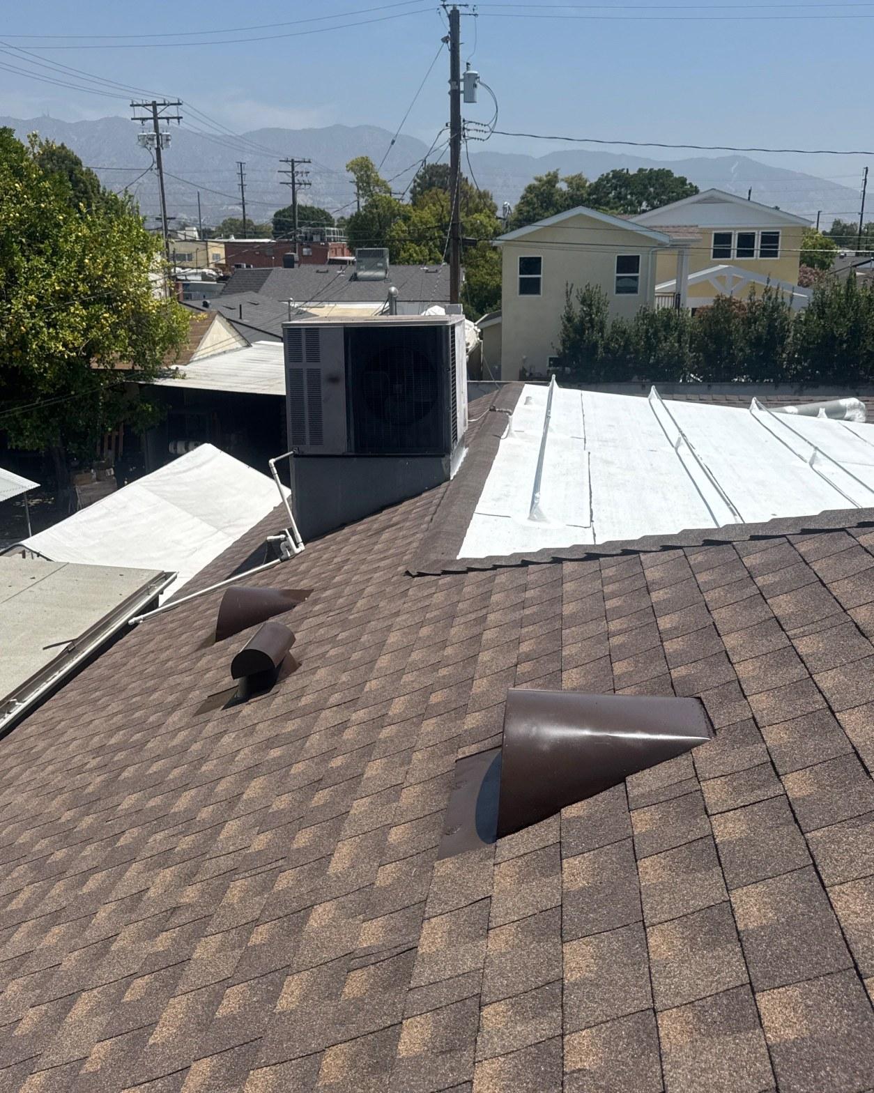 single-reroof-burbank-ca-shingles-flat-flashing-586 Commercial HVAC unit on flat roof next to brown asphalt shingle slope with two pipe vents, suburban LA backdrop.