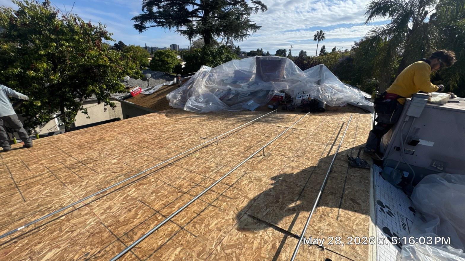 single-reroof-burbank-ca-shingles-flat-flashing-588 Roofers on flat OSB roof deck with solar conduit runs and materials staged under plastic sheeting, Los Angeles suburb.