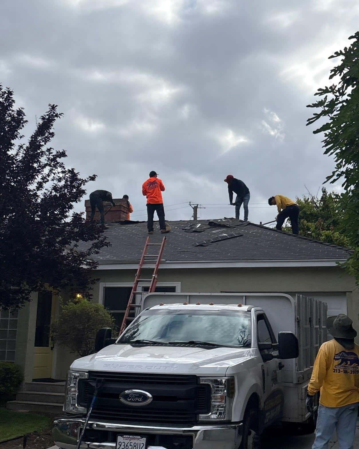 single-reroof-burbank-ca-shingles-flat-flashing-591 Roofing crew tearing off dark asphalt shingles on single-story house with orange ladder and Ford Super Duty work truck.