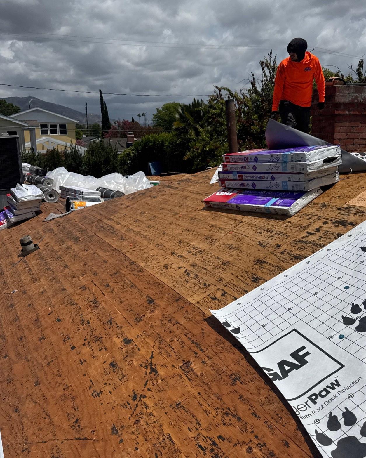 single-reroof-burbank-ca-shingles-flat-flashing-593 Roofer in orange shirt laying GAF WeatherPaw underlayment on bare plywood deck with Timberline HDZ shingle bundles nearby.