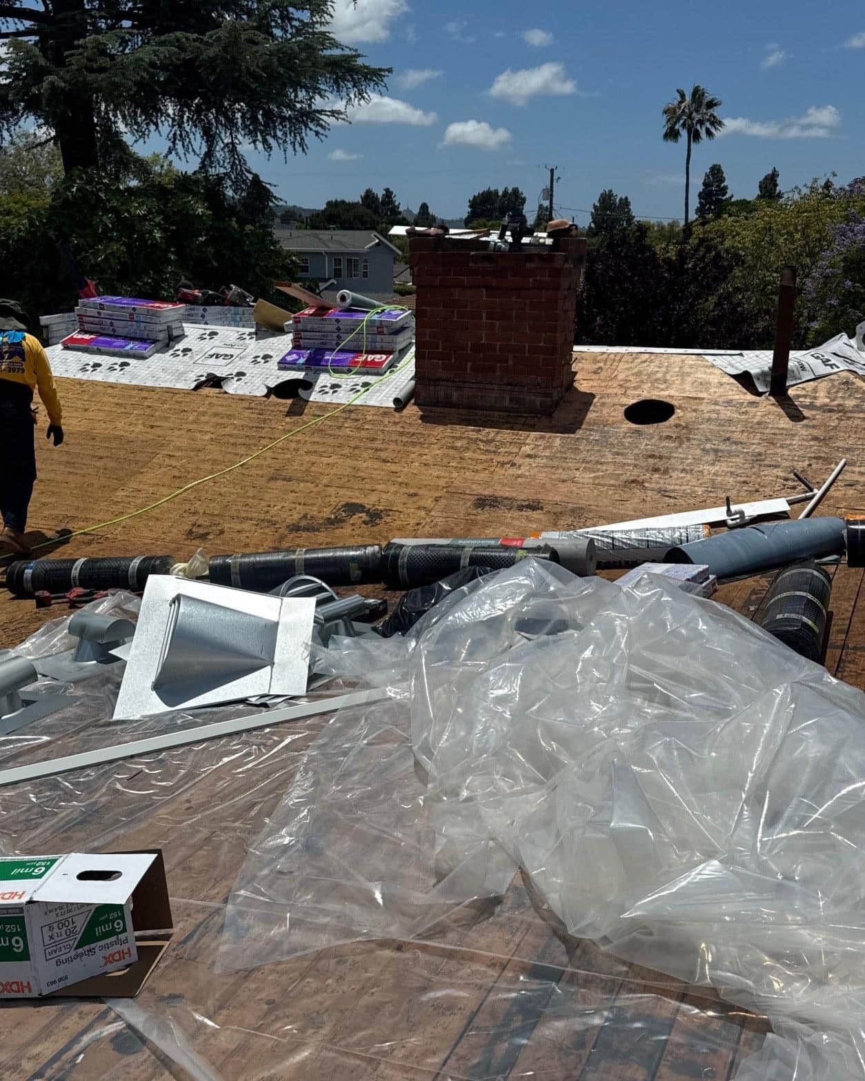 single-reroof-burbank-ca-shingles-flat-flashing-594 Roofing tear-off in progress showing bare plywood deck, GAF underlayment bundles, brick chimney, and metal flashing components on suburban home.