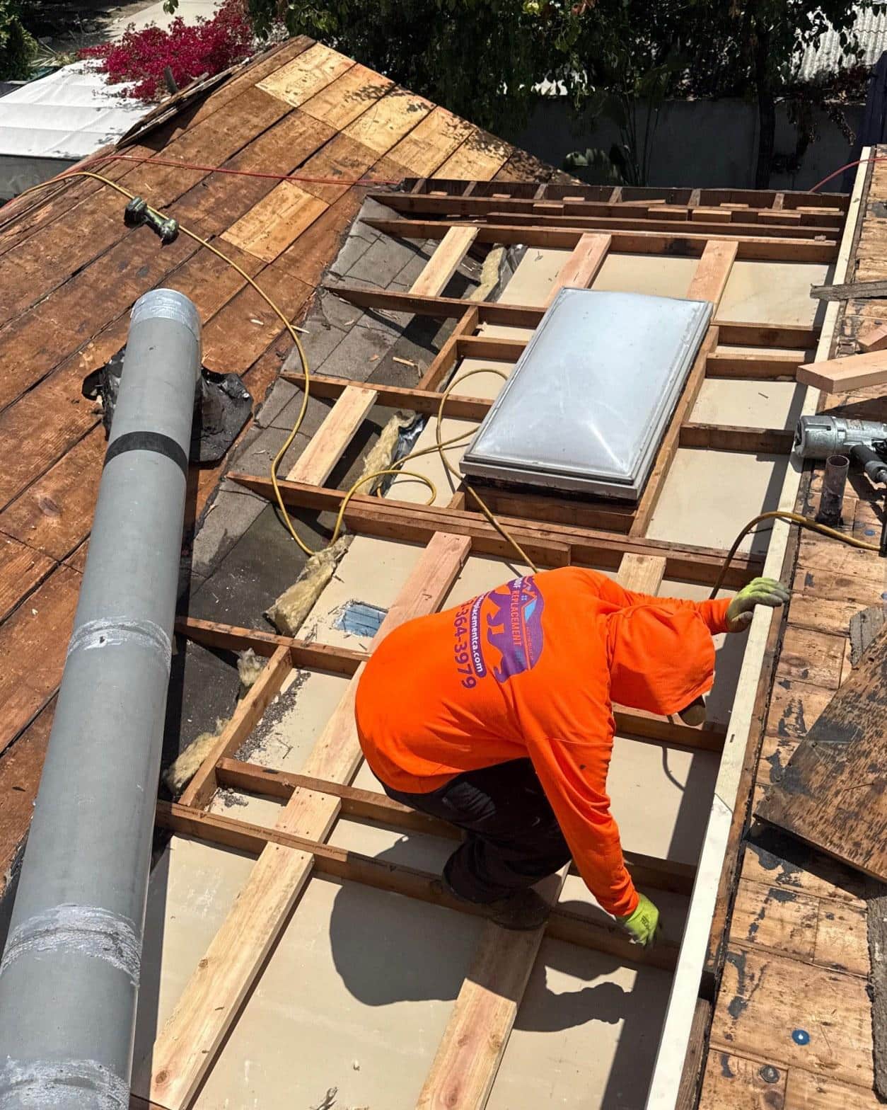 single-reroof-burbank-ca-shingles-flat-flashing-595 Worker in orange shirt installing skylight on stripped roof with exposed rafters and plywood decking.