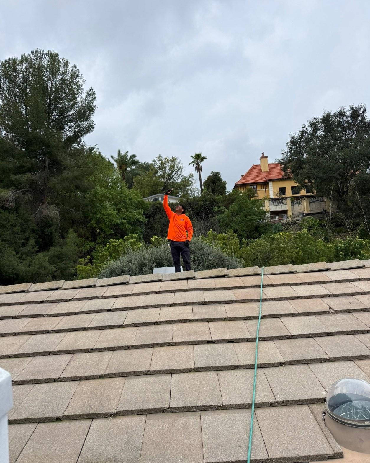 Roofing worker in orange jacket standing on concrete tile roof, pointing upward, with hillside homes in background.