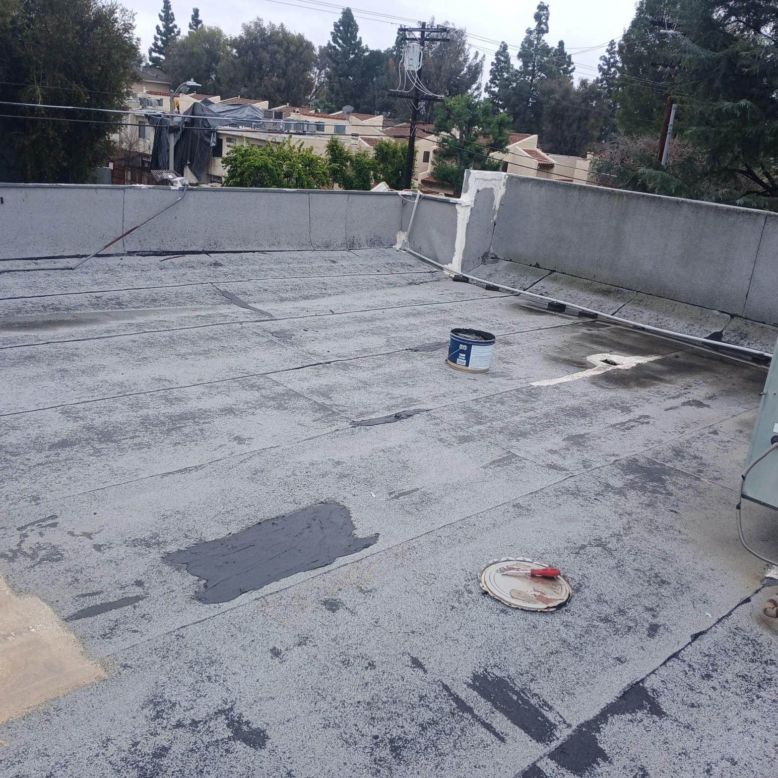 Flat gravel roof with black tar patch repair, open bucket of roofing sealant and brush on rooftop.