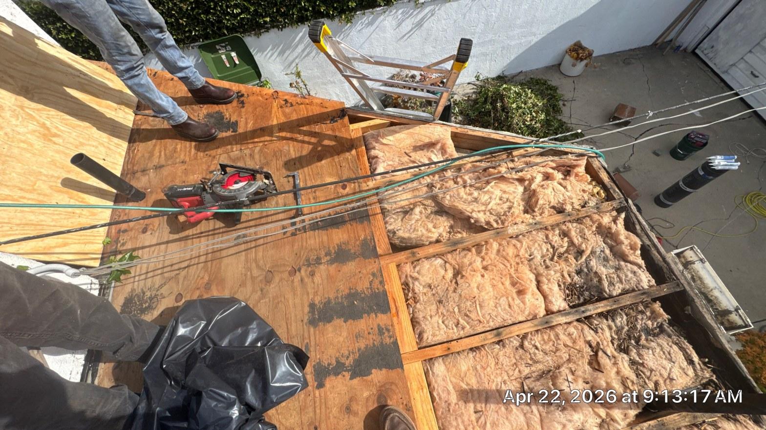 Open roof with plywood sheathing removed, revealing deteriorated fiberglass batt insulation between wood rafters, circular saw on deck.