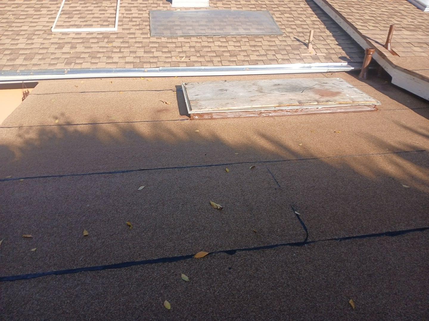 Flat roof covered in rolled roofing material with aged skylight curb next to asphalt shingle roof section.