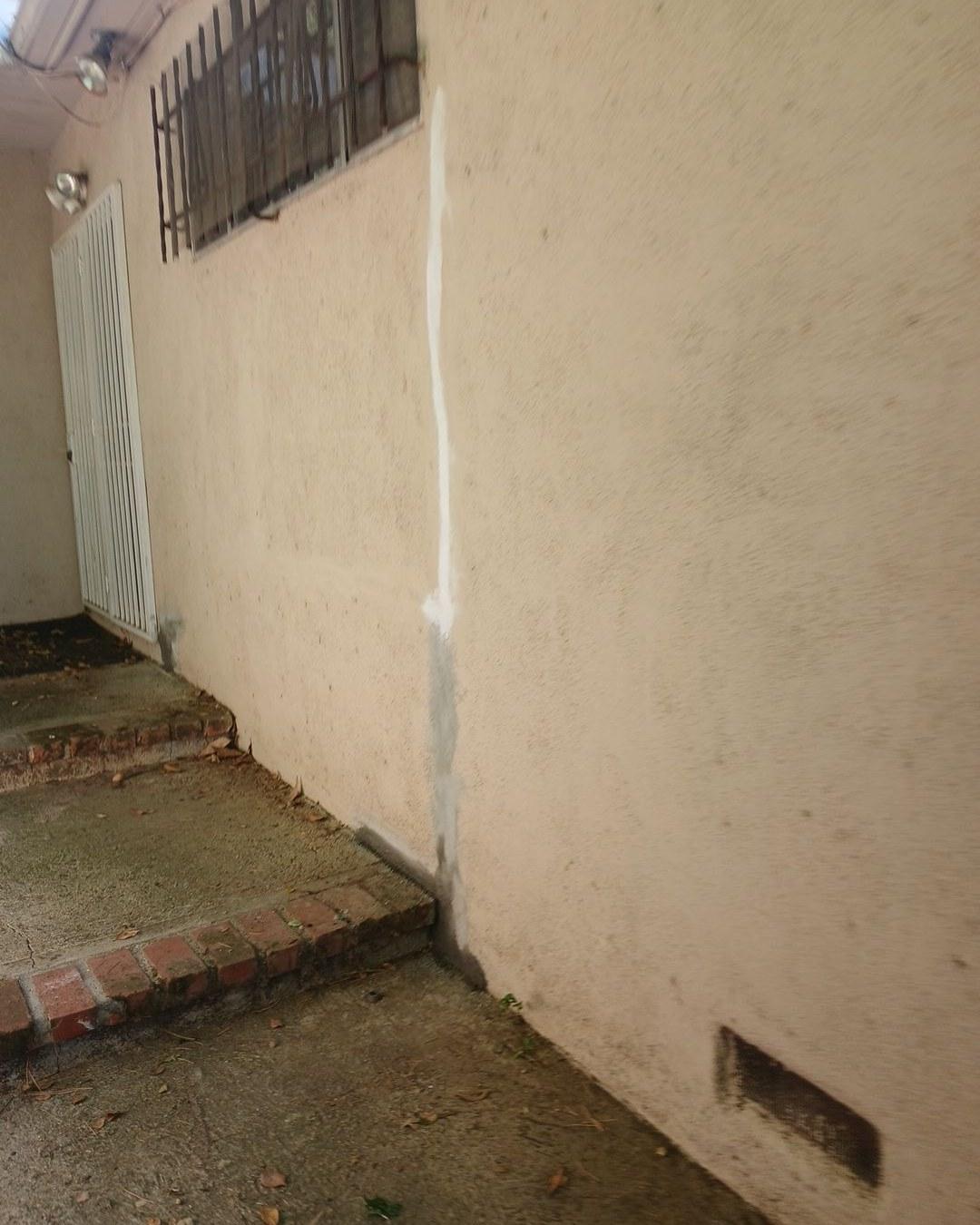 Large vertical crack in beige stucco exterior wall repaired with white caulk sealant near brick pathway.