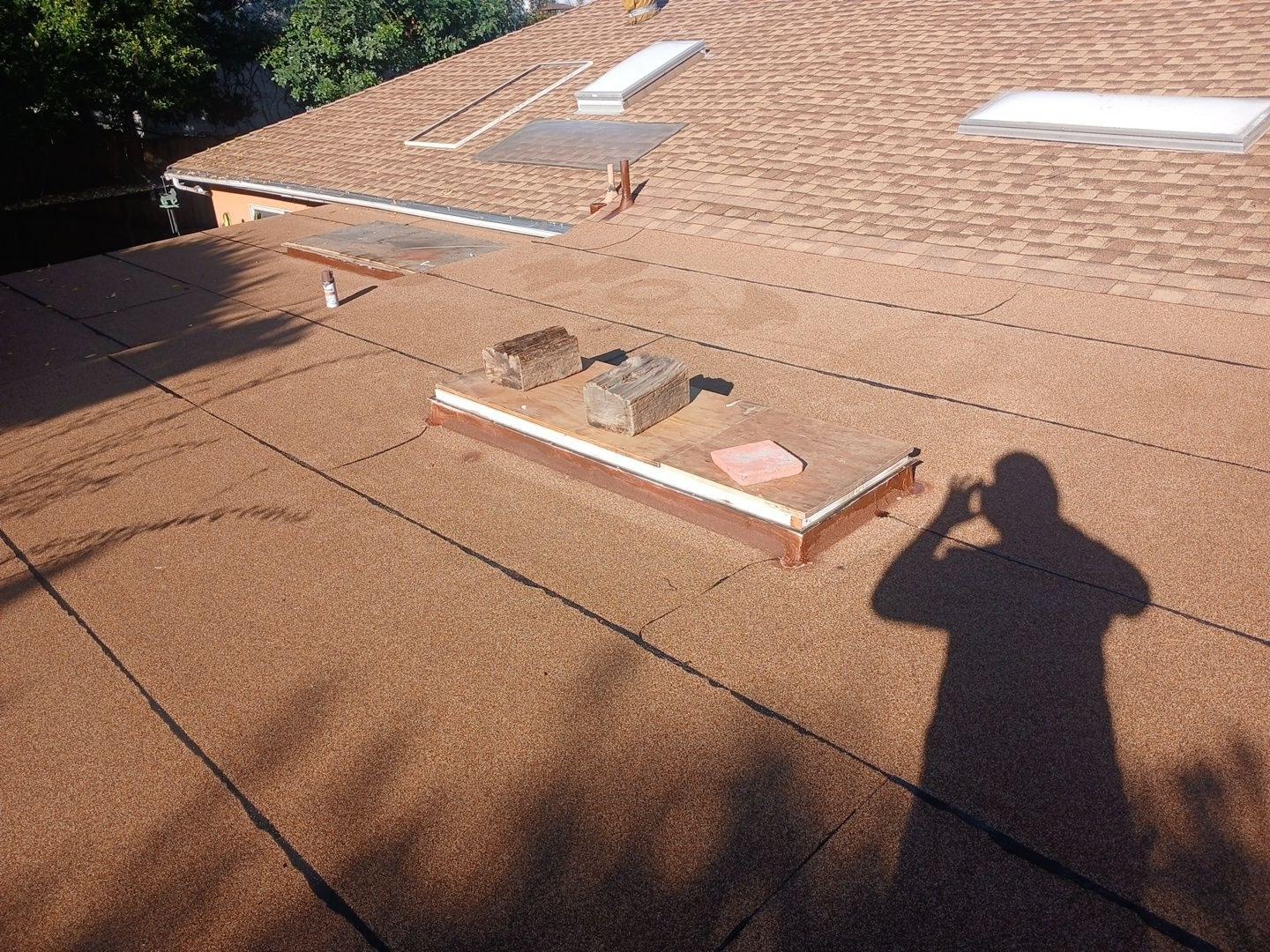 Flat roof with open skylight curb, wood blocks, and asphalt shingle pitched roof section with skylights in background.