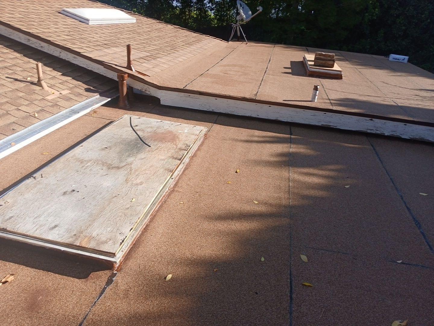 Worn flat roof surface showing open plywood skylight hatch, pipe stacks, parapet edge, and satellite dish.