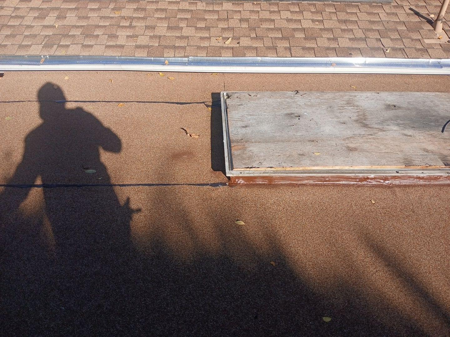 Flat gravel roof with damaged skylight curb, exposed plywood deck, and aluminum flashing near asphalt shingle slope.