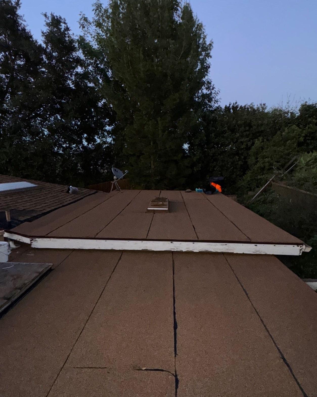 Flat roof covered in brown rolled roofing material with white fascia edge, skylight, and satellite dish in background.
