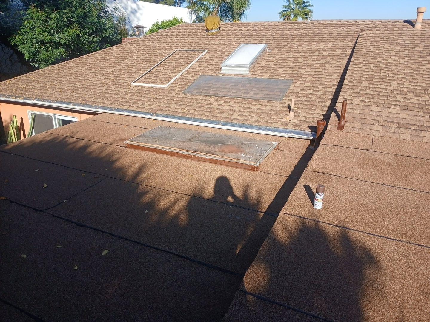 Residential roof with two skylights, flat rolled roofing, brown asphalt shingles, and a spray can sitting nearby.