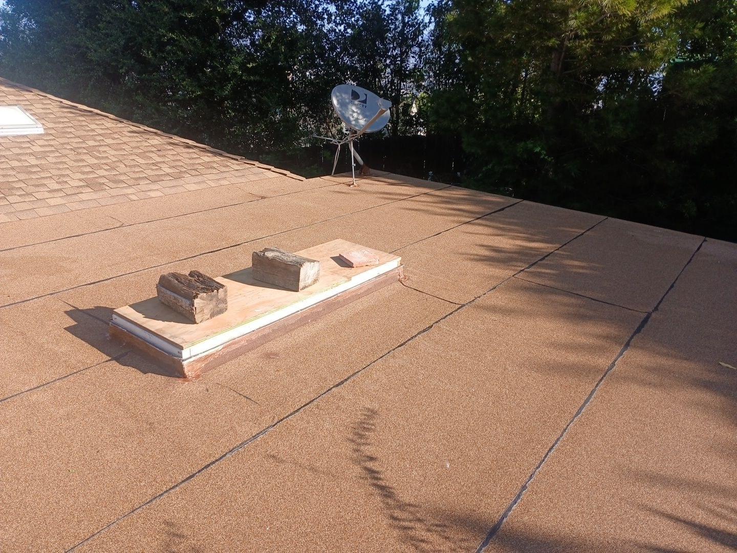 Flat modified bitumen roof with plywood-covered skylight held down by wood blocks and a DirecTV satellite dish in background.
