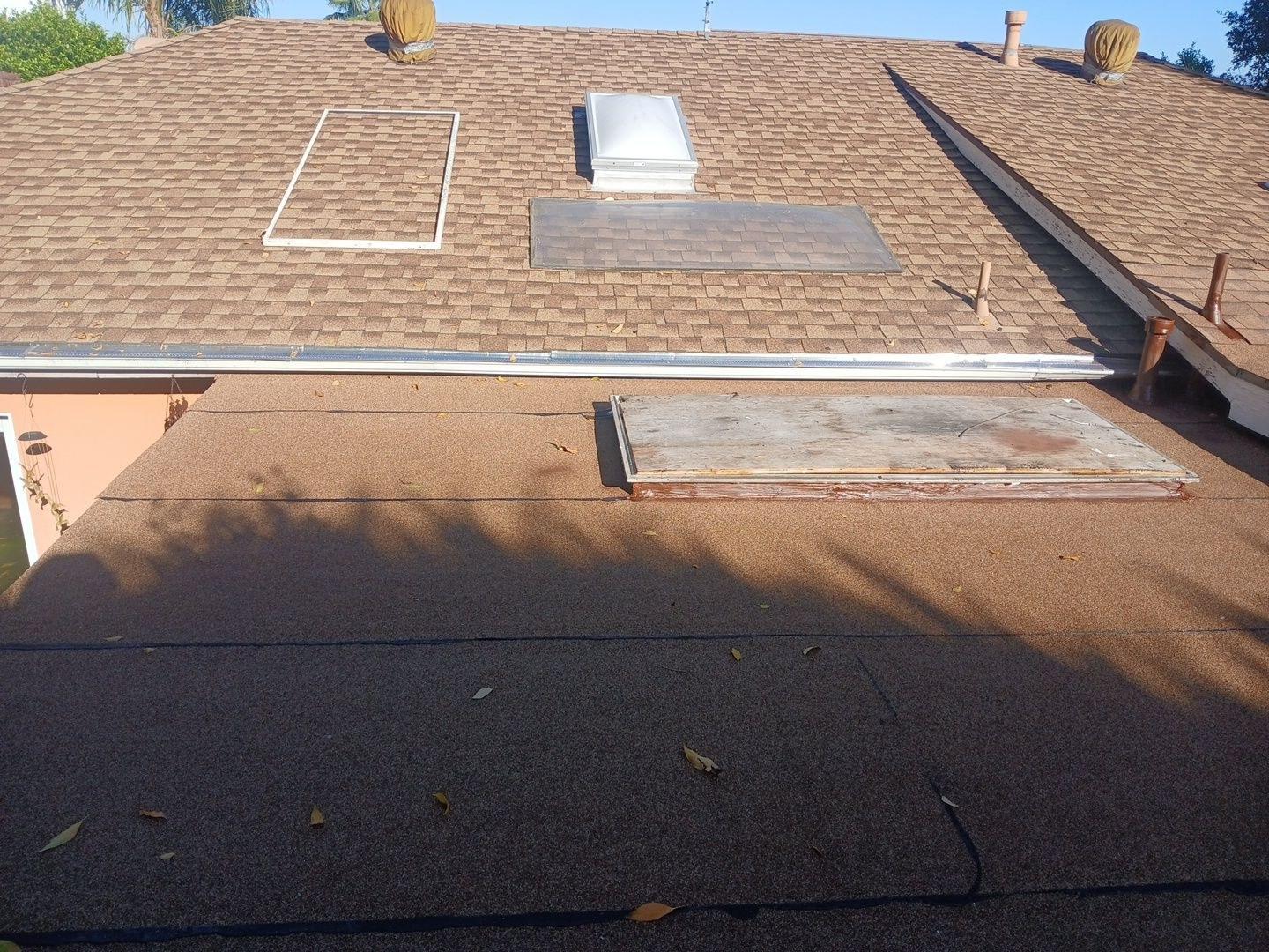 Residential roof showing removed skylight panel, flat rolled roofing, asphalt shingles, and ventilation components in need of repair.