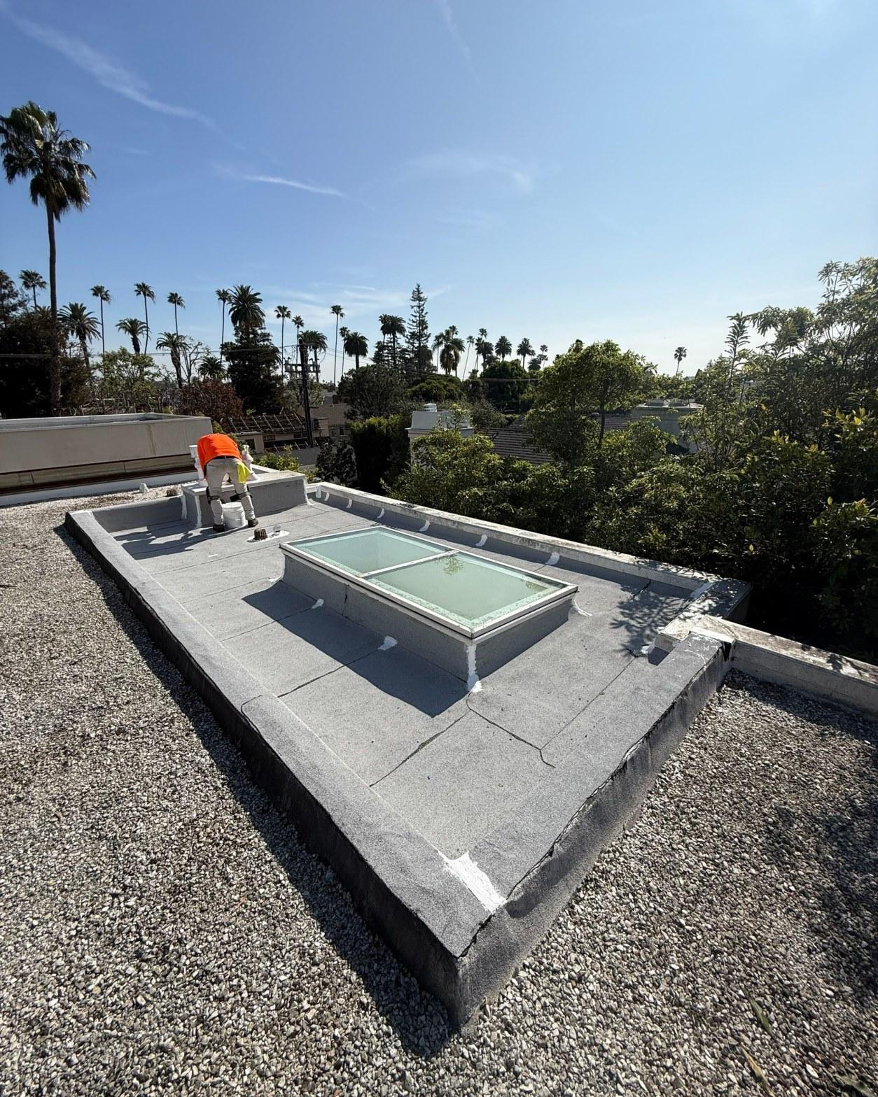 Roofer in orange vest installing glass skylight on flat gravel roof with palm trees in background.
