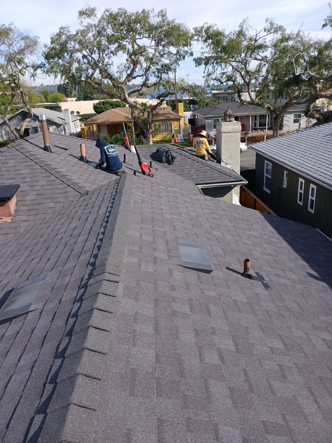 Crew installing dark asphalt shingles on large hip roof with skylights and pipe flashing | Roof Replacement Inc Crew installing dark asphalt shingles on large hip roof with skylights and pipe flashing — Roof Replacement Inc