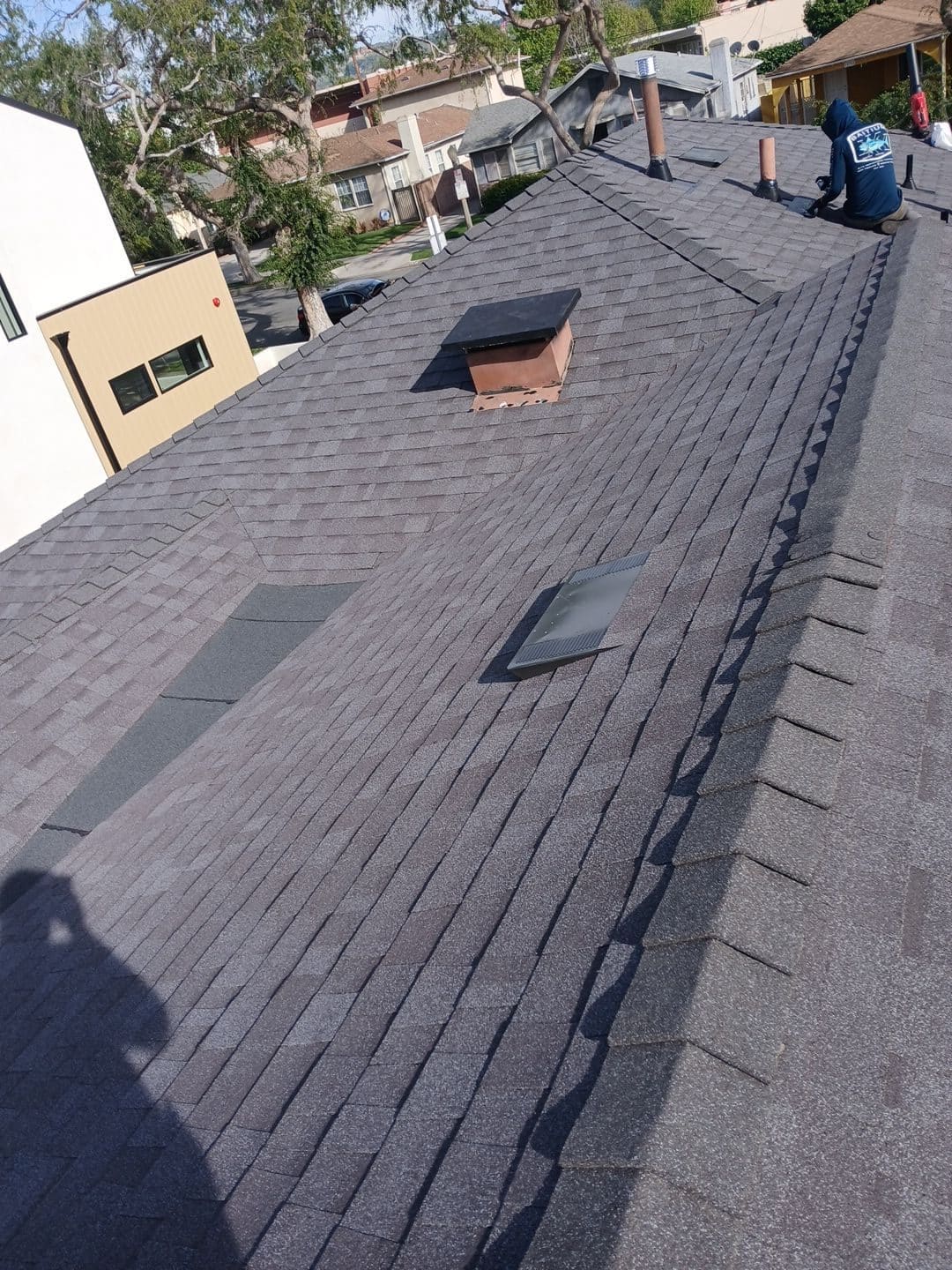 Completed gray asphalt shingle roof with skylight, chimney, vents, worker in background | Roof Replacement Inc Completed gray asphalt shingle roof with skylight, chimney, vents, worker in background — Roof Replacement Inc