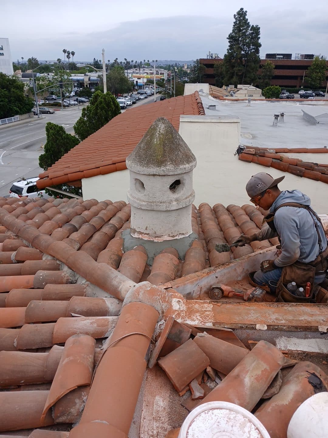 Worker repairing clay barrel tile roof around decorative concrete chimney pot | Roof Replacement Inc Worker repairing clay barrel tile roof around decorative concrete chimney pot — Roof Replacement Inc