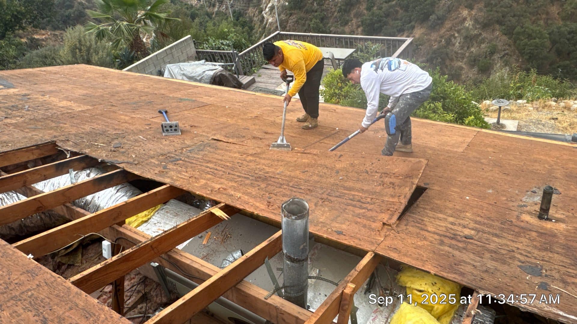 Two roofers removing plywood decking with pry bars, exposed rafters below in Altadena | Roof Replacement Inc two roofers removing plywood decking with pry bars, exposed rafters below — Altadena — Roof Replacement Inc