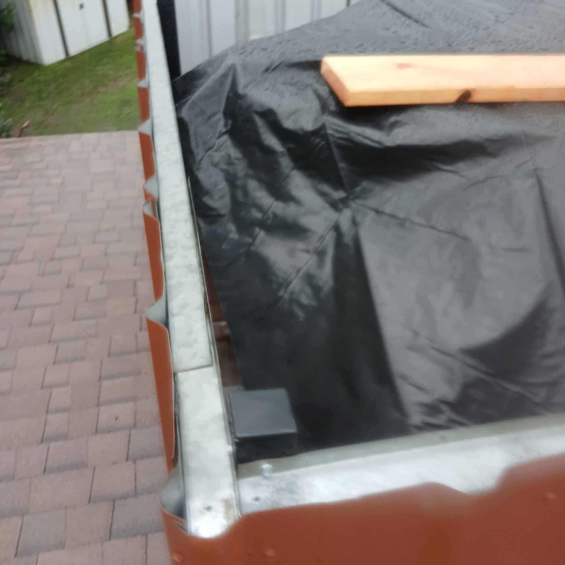 Black poly tarp over roof deck, metal gutter, wood plank weight — Roof Replacement Inc