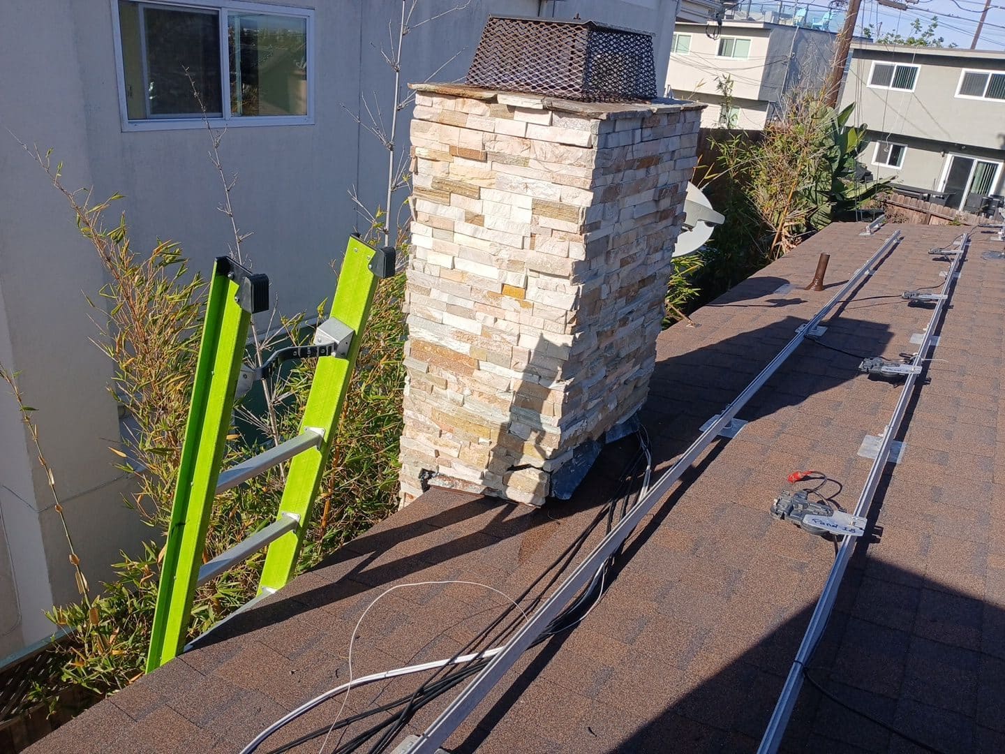 Stone chimney on asphalt shingle roof with solar rail mounting hardware and ladder — Roof Replacement Inc