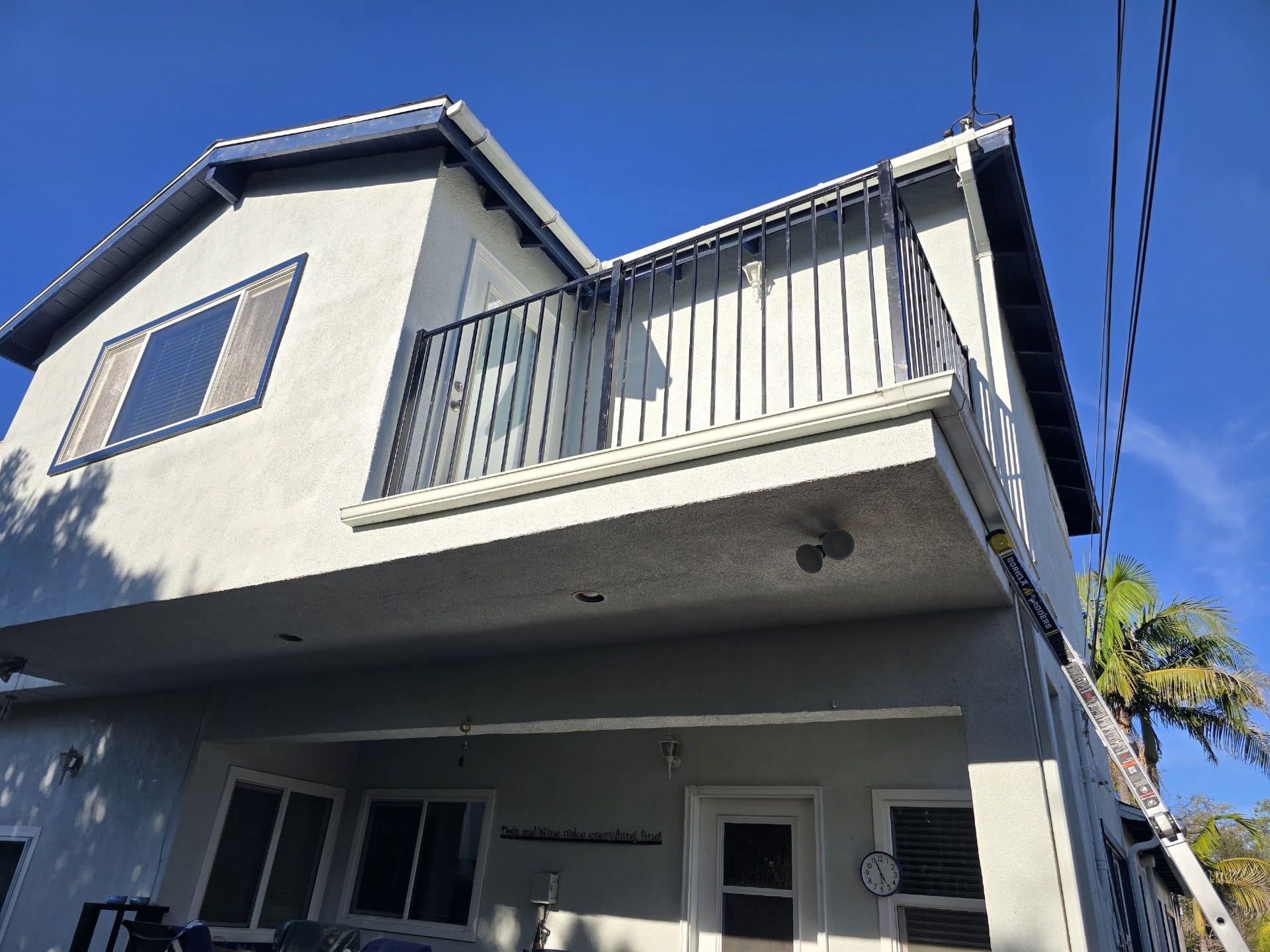 Two-story stucco home, metal balcony railing, gutters, ladder leaning on eave | Roof Replacement Inc Two-story stucco home, metal balcony railing, gutters, ladder leaning on eave — Roof Replacement Inc