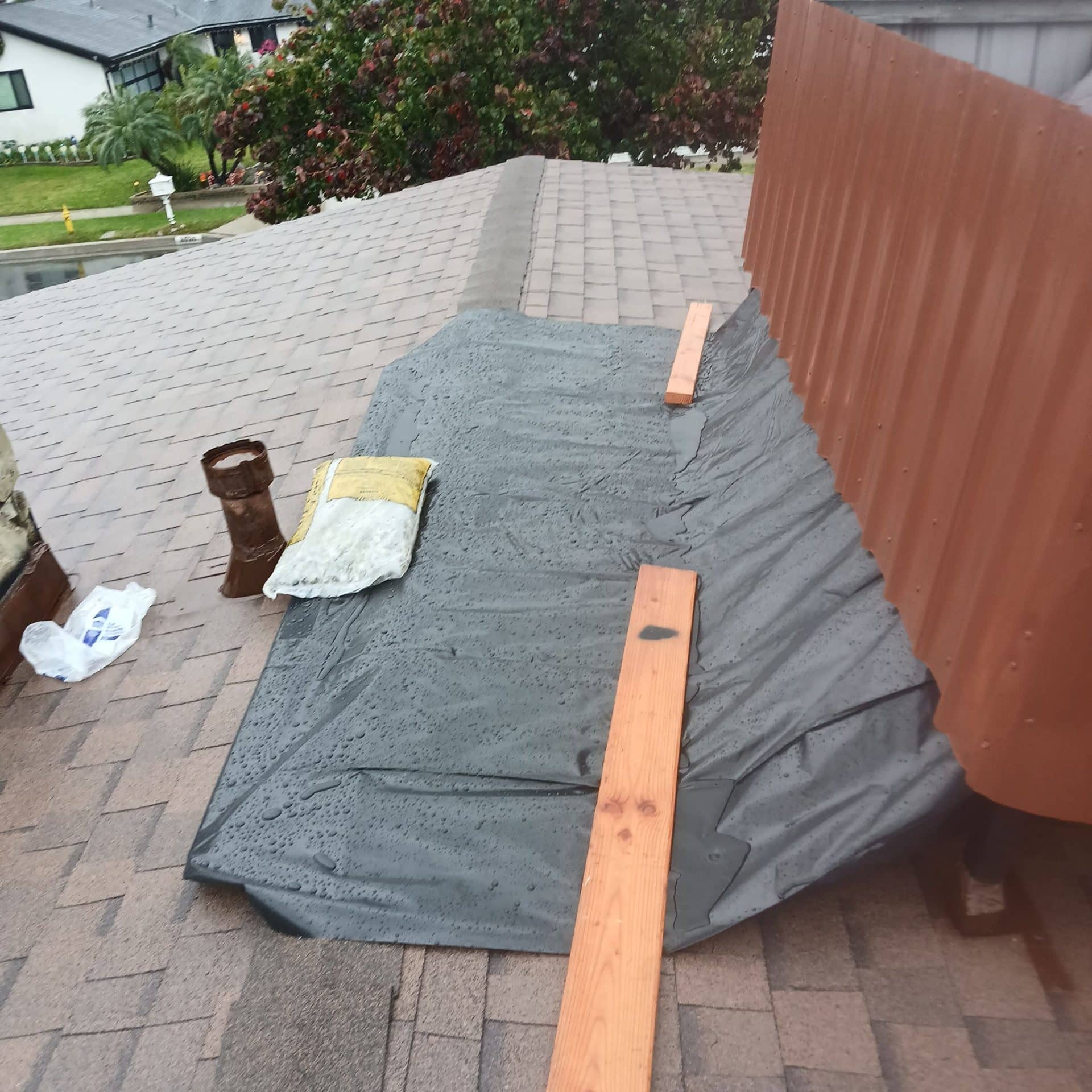 Wet tarp covering shingle roof section beside corrugated metal wall — Roof Replacement Inc