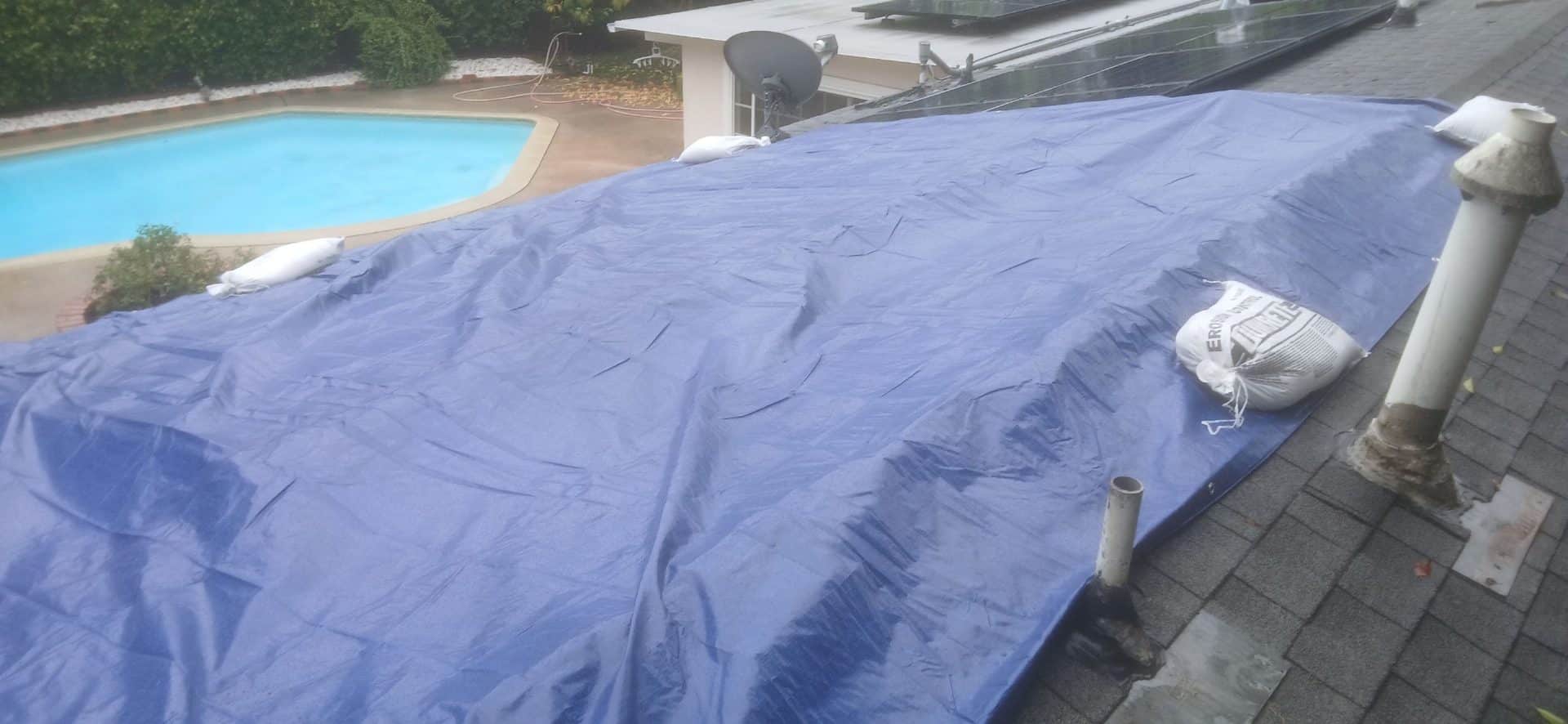 Blue tarp with sandbags covering flat roof section, pipe penetrations visible — West Hills — Roof Replacement Inc
