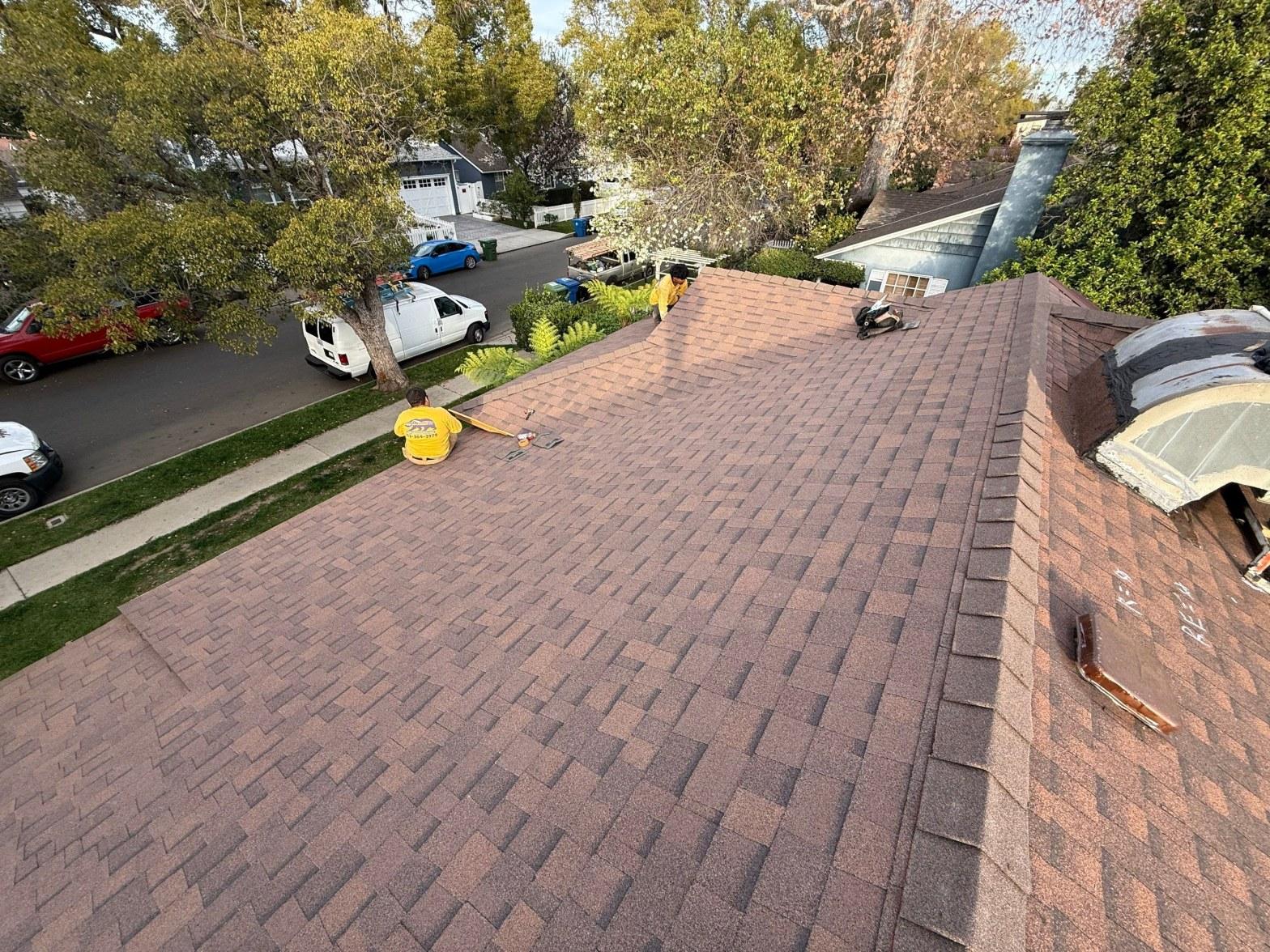 Two workers in yellow shirts laying red asphalt shingles on a residential home roof in a suburban neighborhood.