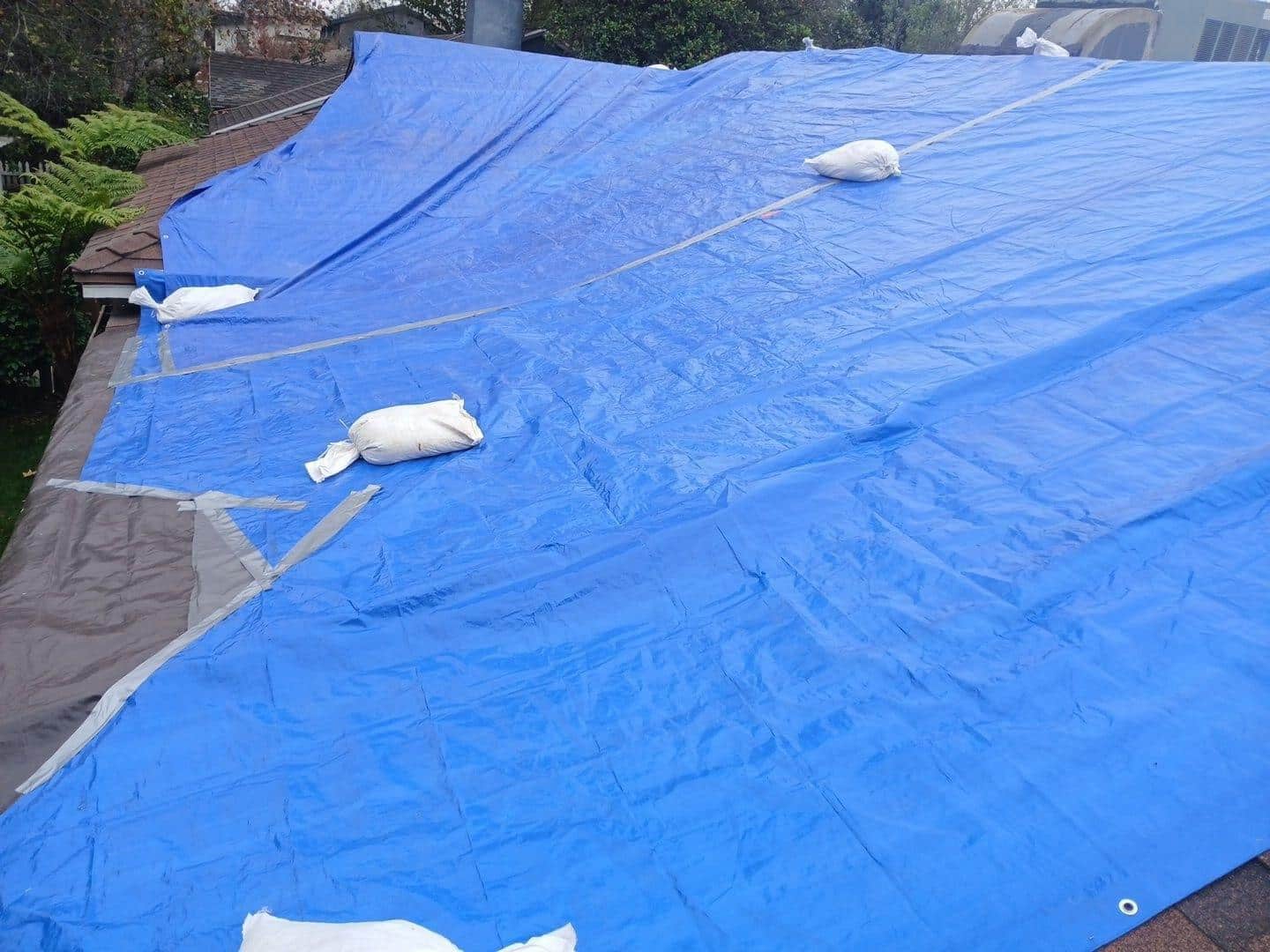 shingle-reroof-duration-cool-plus-los-angeles-3313 Blue polyethylene tarps weighted down with white sandbags covering a storm-damaged residential roof.