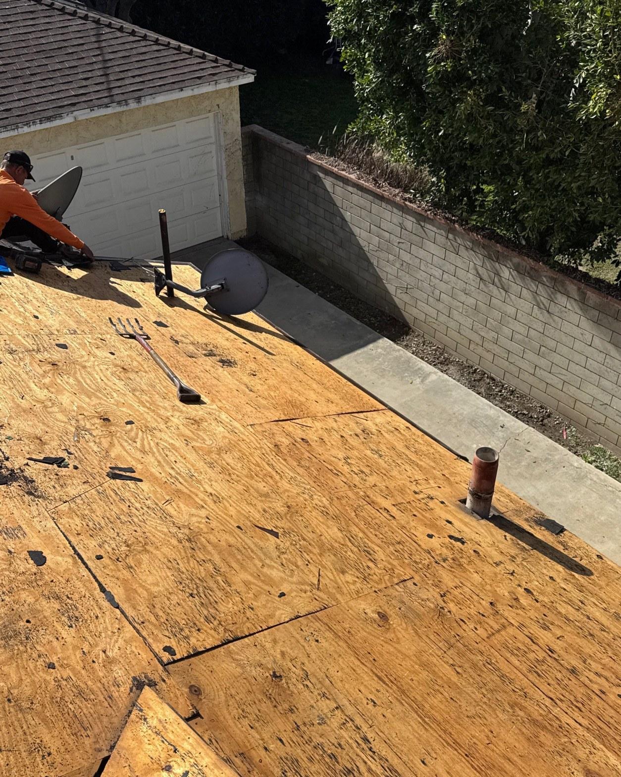 shingle-reroof-duration-cool-plus-los-angeles-3315 Roofing contractor in orange shirt working on bare plywood roof deck with pipe penetration and satellite dish visible.