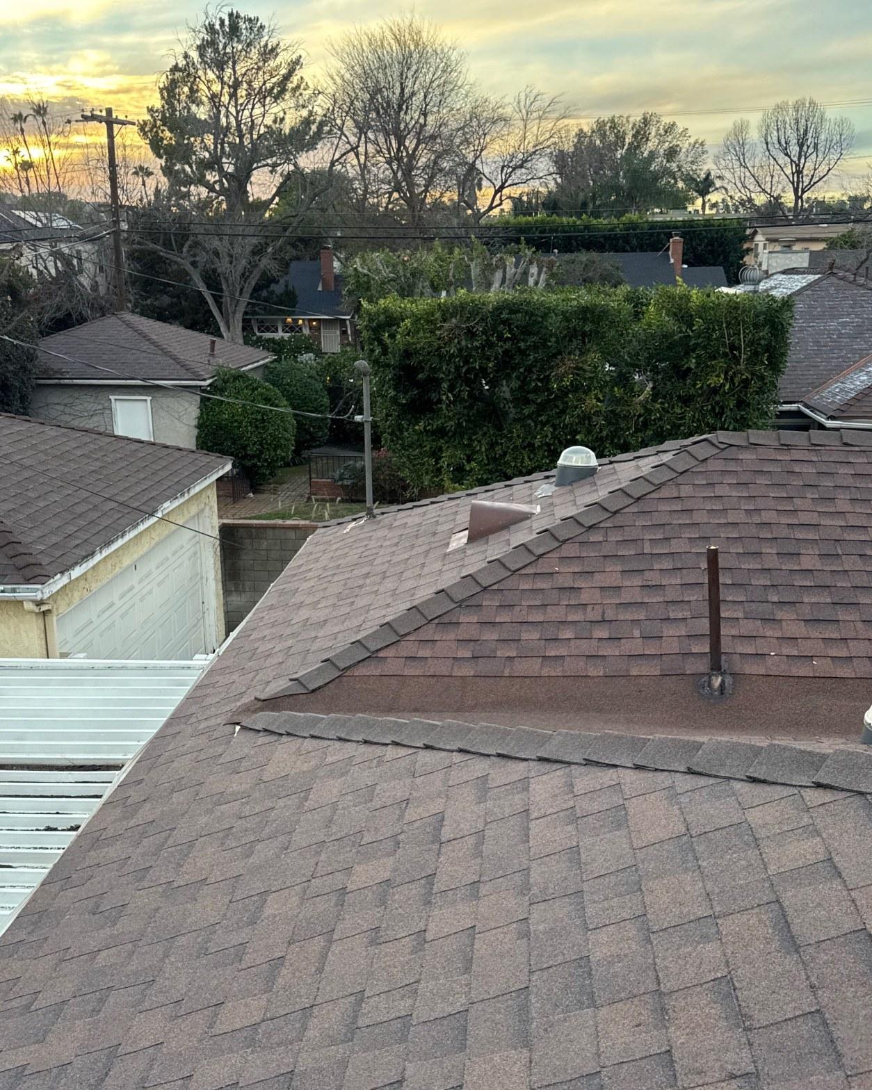 shingle-reroof-duration-cool-plus-los-angeles-3323 Residential brown asphalt shingle roof with plumbing stack pipe and dome vent, suburban neighborhood at sunset.