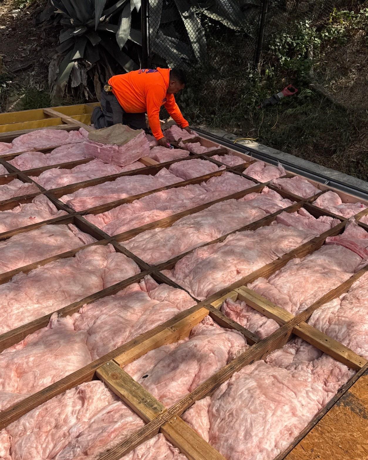 Worker fitting pink fiberglass batt insulation into roof joist bays before new decking installation