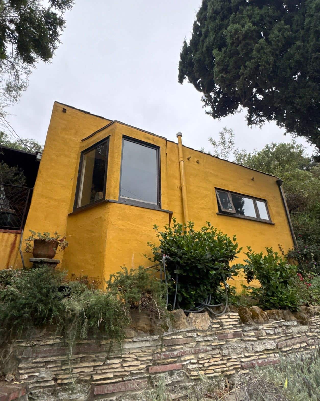 torch-down-flat-roof-replacement-insulation-los-angeles-3337 Bright yellow stucco exterior of small building with dark-framed windows, stone retaining wall, and surrounding shrubs.