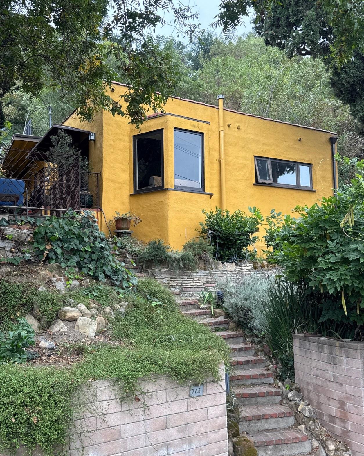 torch-down-flat-roof-replacement-insulation-los-angeles-3338 Bright yellow stucco home with dark-framed windows, brick stairway, and lush hillside landscaping, address 713.