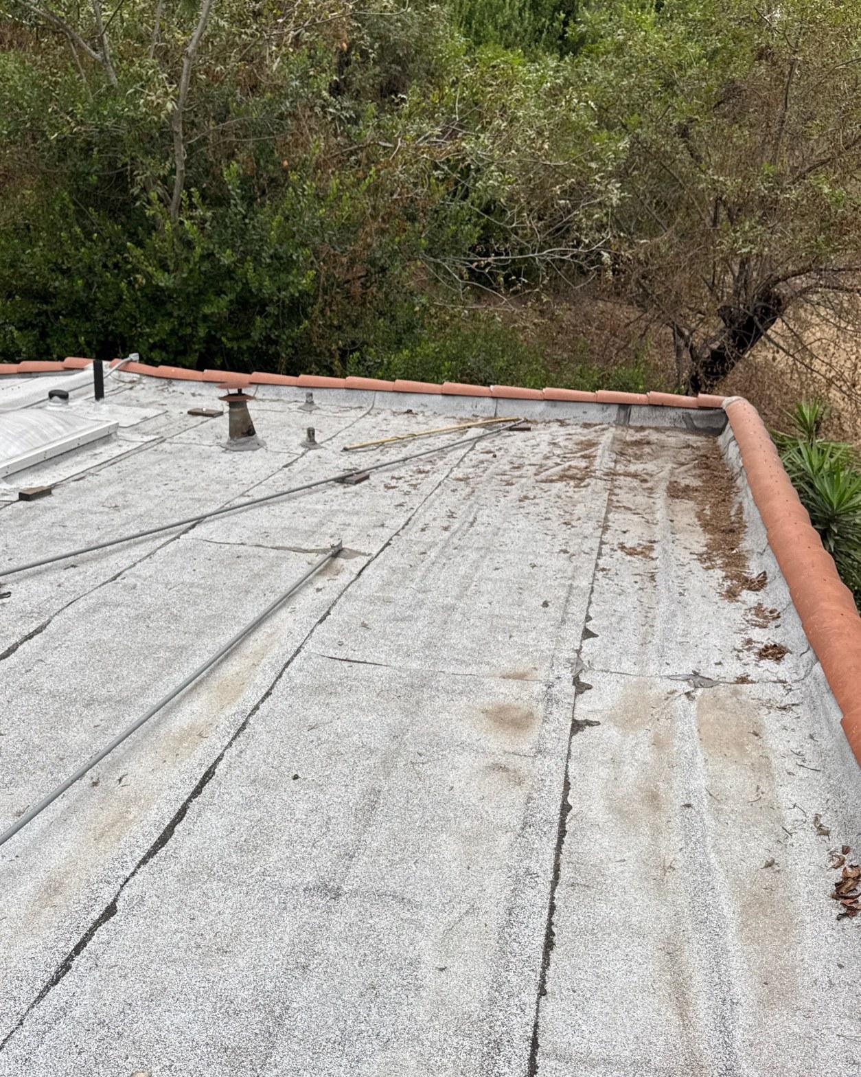 torch-down-flat-roof-replacement-insulation-los-angeles-3342 Worn flat roof surface showing cracks, dirt buildup, and clay tile parapet edging with trees in background.